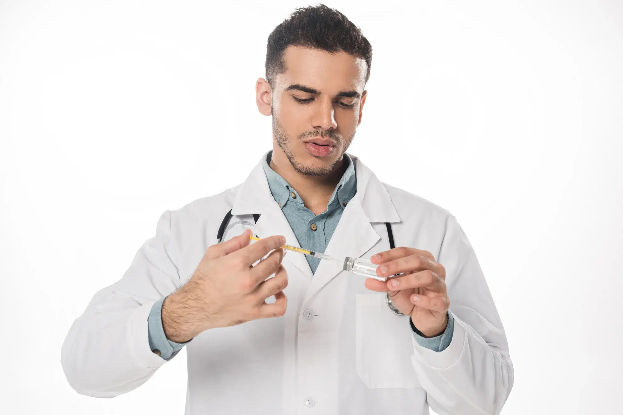A male doctor in a white lab coat prepares a syringe by drawing liquid from a vial, highlighting his expertise in administering treatments like Botox for the scrotum. He is focused on his task and stands against a plain white background, with a stethoscope draped around his neck.