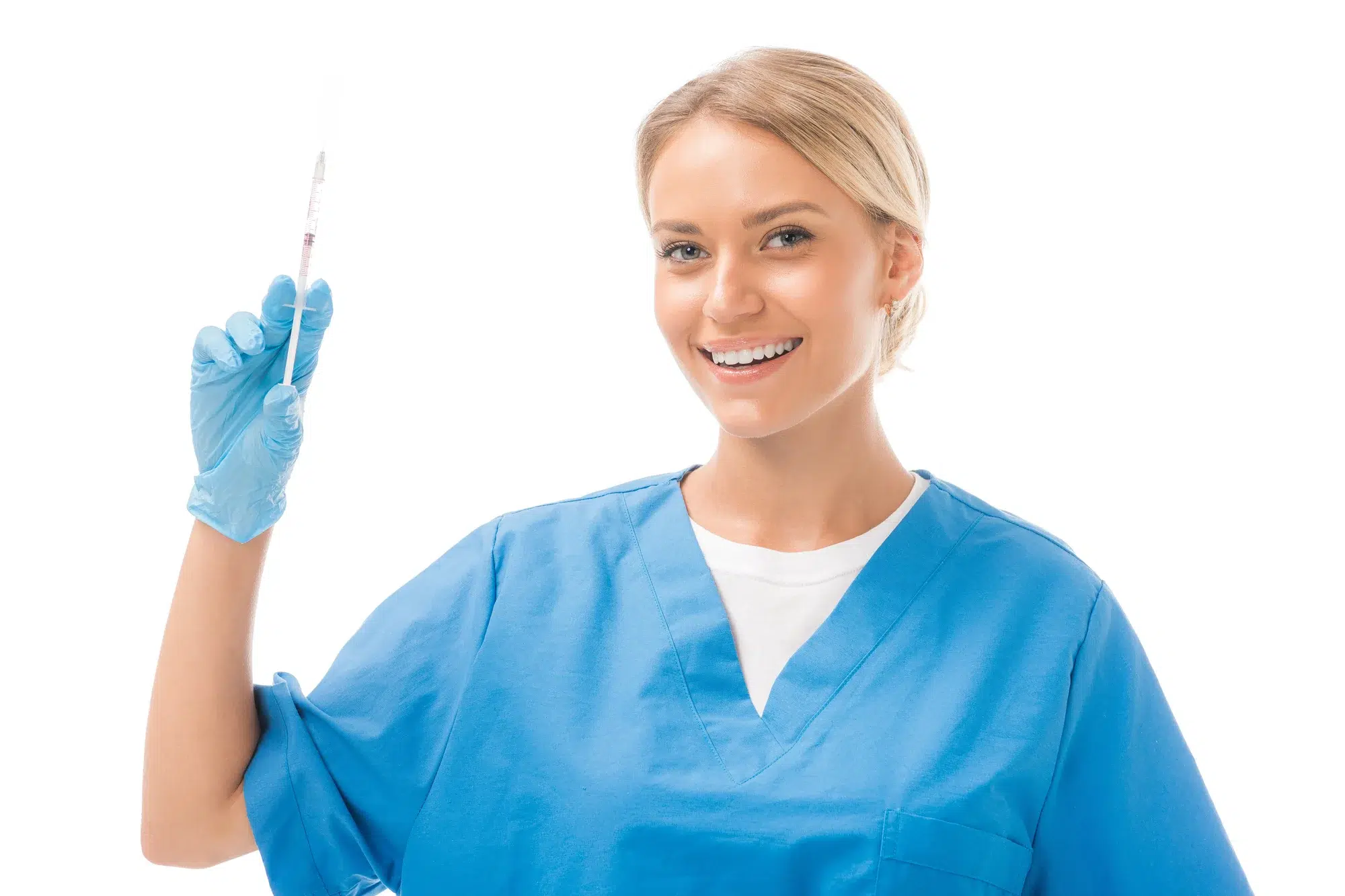A healthcare professional wearing blue scrubs and blue gloves is smiling and holding a syringe upright in one hand. She has blonde hair pulled back and stands against a white background, ready to administer Juvederm for penis enhancement.