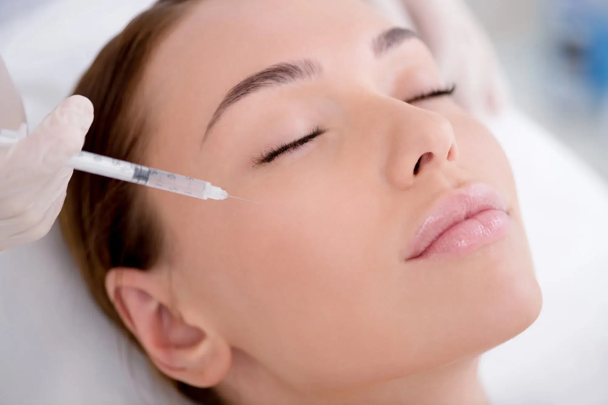 A woman with closed eyes is lying down while a gloved hand holds a syringe and prepares to administer an injection to the skin near her eye. The scene appears to be in a clinical or spa setting. Her expression is calm and serene.