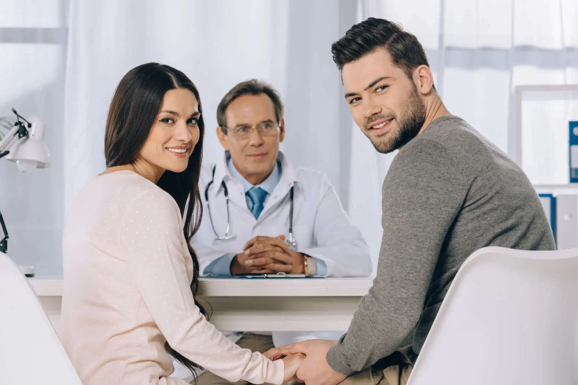 A smiling couple holds hands while seated at a desk with a doctor in a white coat. The doctor, specializing in Botox for Incontinence, sits opposite them with hands clasped, as they discuss treatment options in a bright, well-lit medical office.