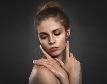 A young person with light skin and long dark hair tied in a bun poses against a dark background. With eyes closed and a serene expression, they gracefully position their hands under their chin and by their cheek, embodying the effortless beauty often sought by those who choose the best dermal fillers (top brands).