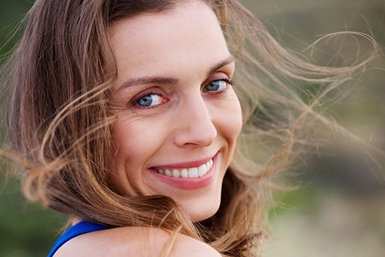 A woman with long, brown hair smiles at the camera as the wind blows her hair across her face. She is wearing a blue top, and her light-colored eyes are accentuated by natural lighting. The blurred background suggests an outdoor setting. Those wondering "How Long Does Juvederm Voluma Last" might notice her youthful appearance shines through naturally.