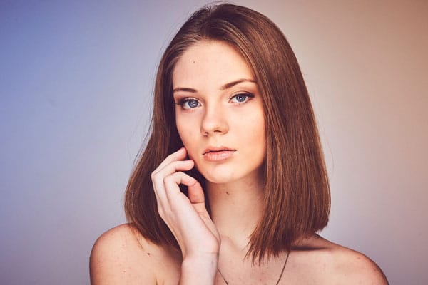 A young person with straight, shoulder-length brown hair and light blue eyes poses against a gradient background that transitions from blue to beige. They have a calm expression, with one hand gently touching their face.