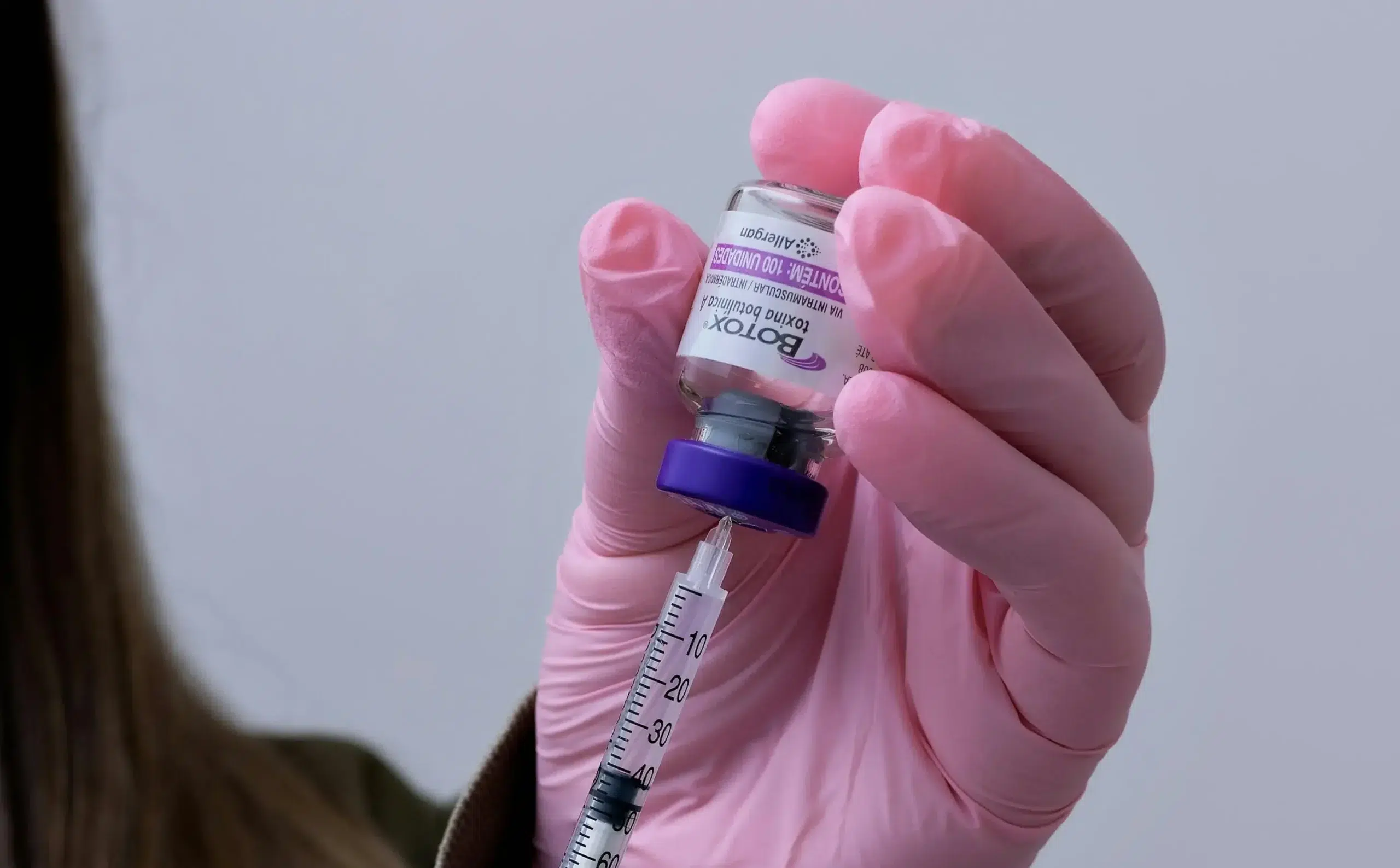 A person wearing pink gloves holds a small vial labeled "Botox," drawing the substance into a syringe. The background is blurred and neutral in color, subtly hinting at its intriguing use—Botox for the penis.