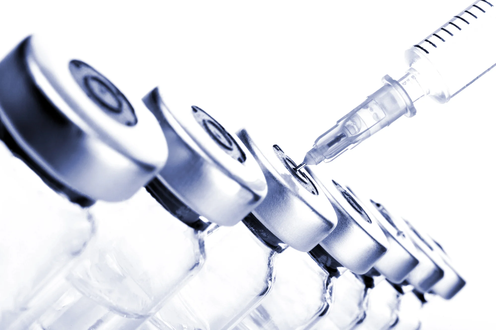 Close-up image of several glass vials with metal caps arranged in a row. A syringe is placed above one of the vials, with its needle piercing the vial's cap. The clinical scene highlights neurotoxins, depicted with a sterile appearance and high-key lighting effect.