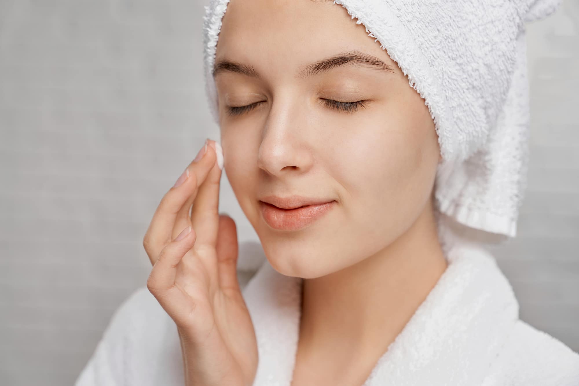 Woman with closed eyes applying moisturizer on face.