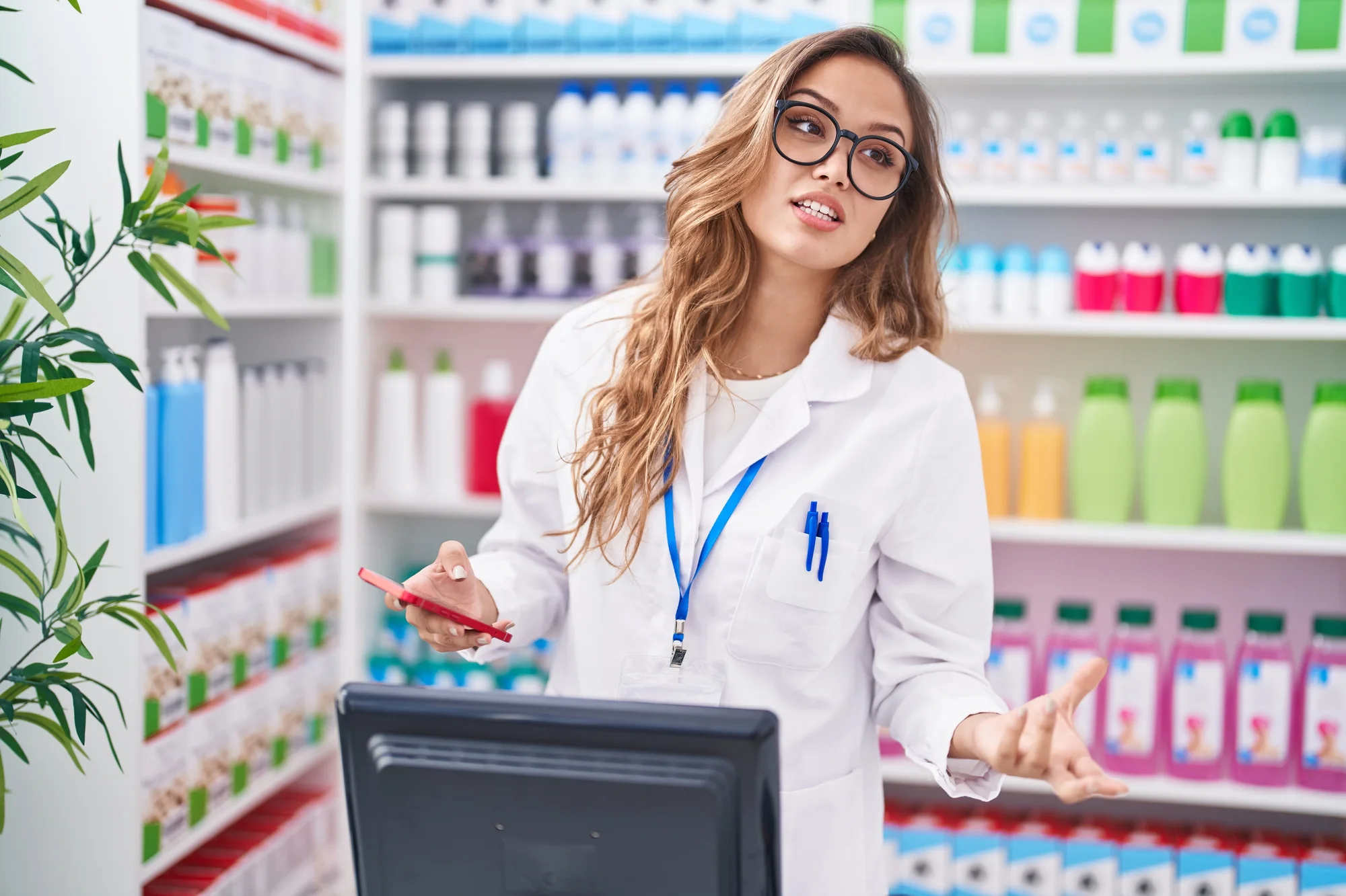 A woman in a white lab coat stands behind a counter, speaking and holding a red phone. She has long hair, glasses, and a name badge. Shelves stocked with various colorful bottles and products are visible in the background. A potted plant is on the left.