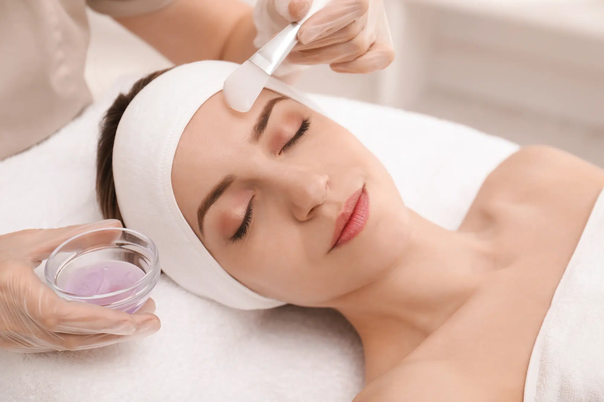 A woman with a white headband is lying down with her eyes closed. A person wearing gloves is applying a facial mask on her forehead using a spatula. A small bowl of purple facial mask product is held in one hand. The setting appears to be a spa.