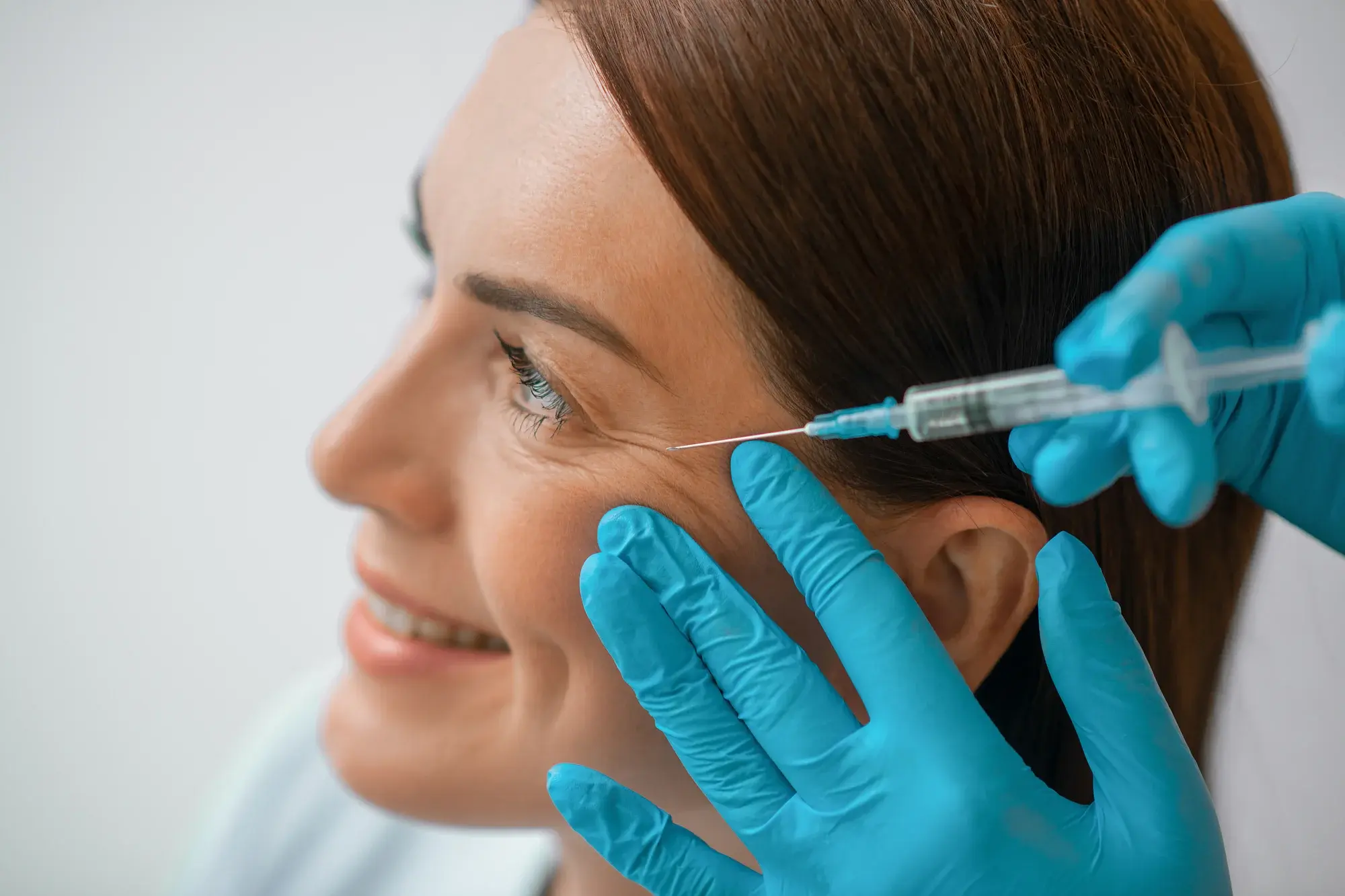 A close-up of a woman smiling as a gloved hand administers an injection near her right eye area. The woman has smooth skin and straight brown hair, and the gloved hands are wearing blue medical gloves.