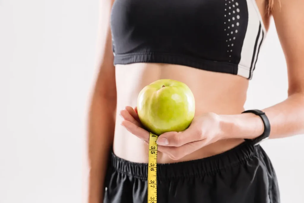 An individual wearing their workout clothes, holding an apple and tape measure.