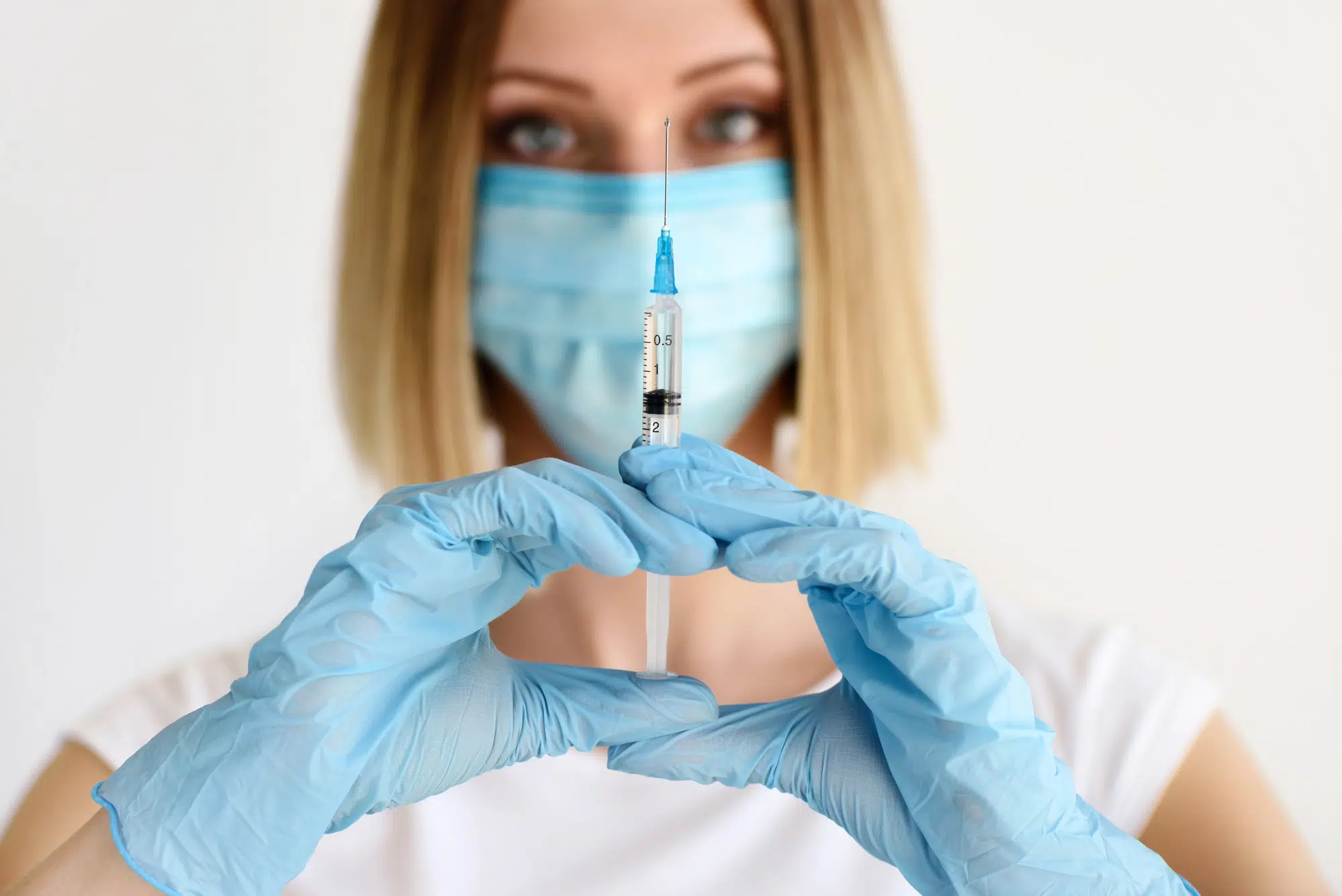 A person wearing a blue mask and gloves holds a syringe vertically. The focus is on the syringe, which is in the center of the image. The background is a plain, light color.