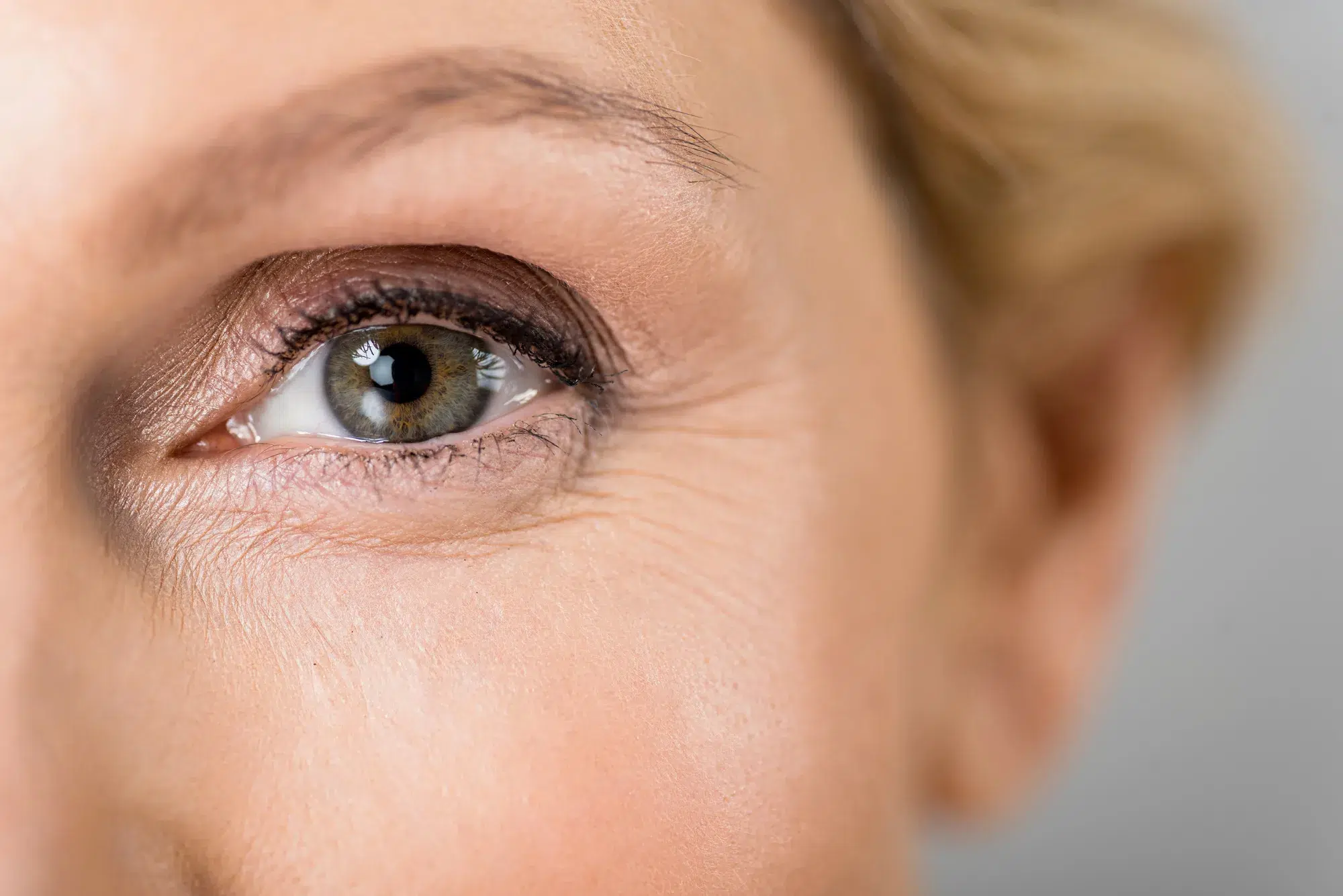 Close-up of a person's eye, highlighting the details such as the eyelashes, light makeup, and the natural texture of the surrounding skin. The person has fair skin and the eye color appears to be hazel.