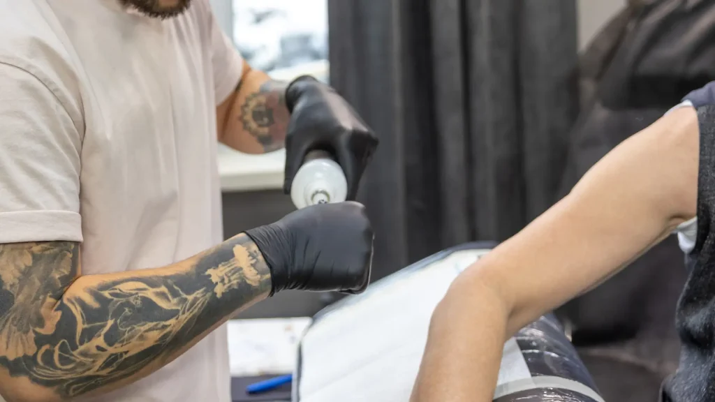 A tattoo artist preparing to apply a numbing cream on client's skin.