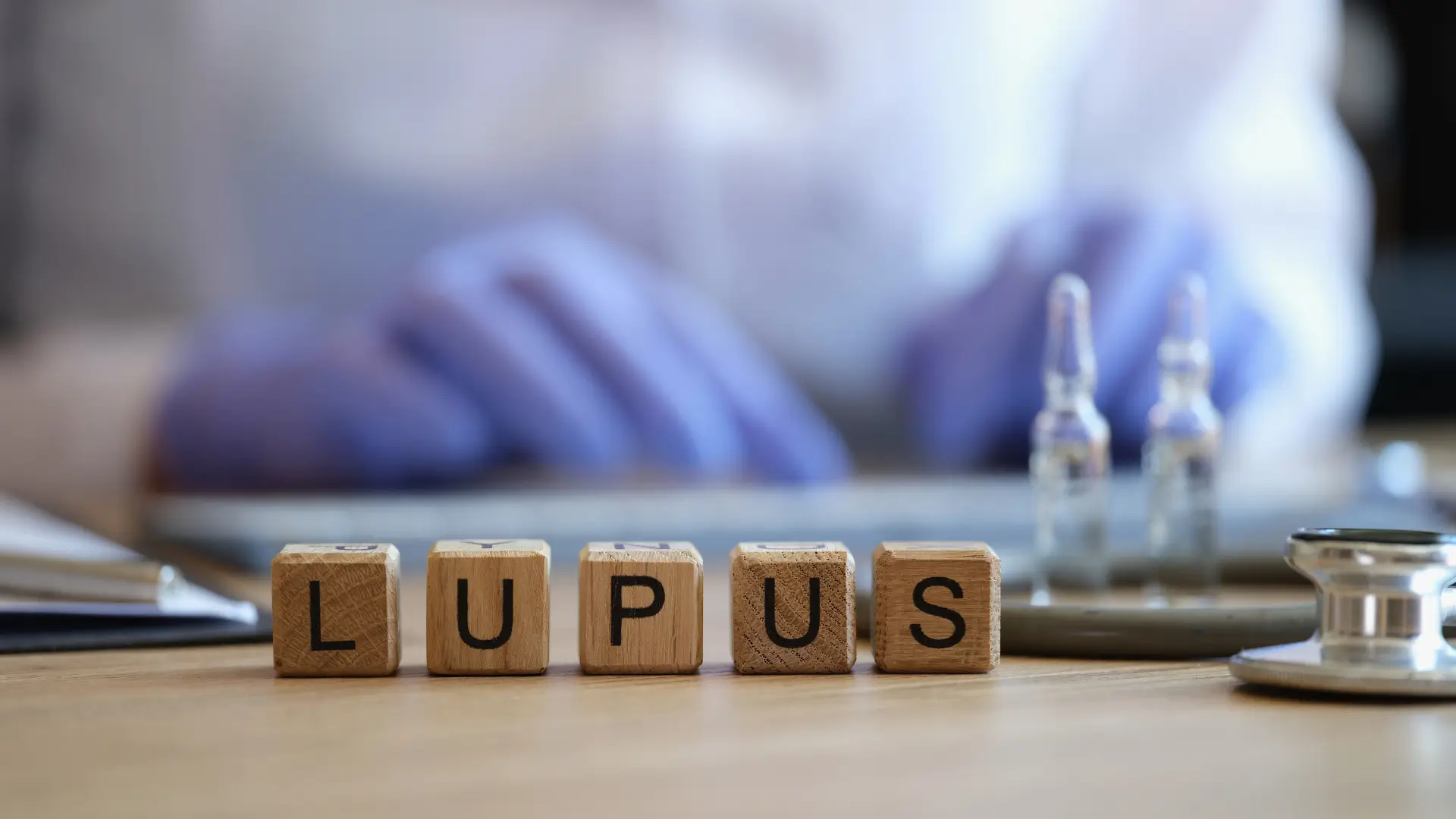 A close-up shot of blocks that spelled lupus with a blurry photograph of a doctor at the back.